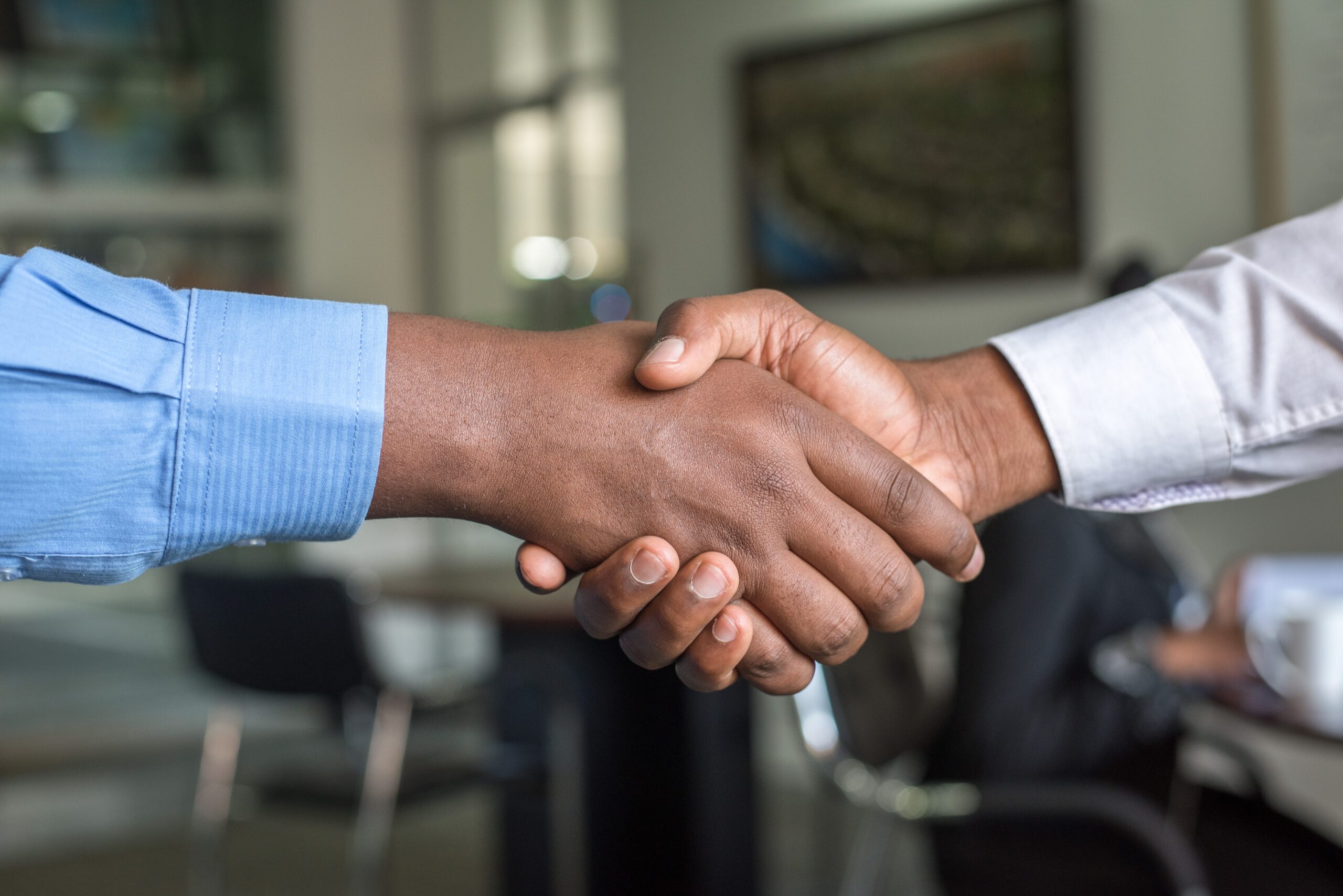 Two people shaking hands in an office setting, with focus on their hands and a blurred background featuring chairs and tables, signify the crucial role of trust between executors.