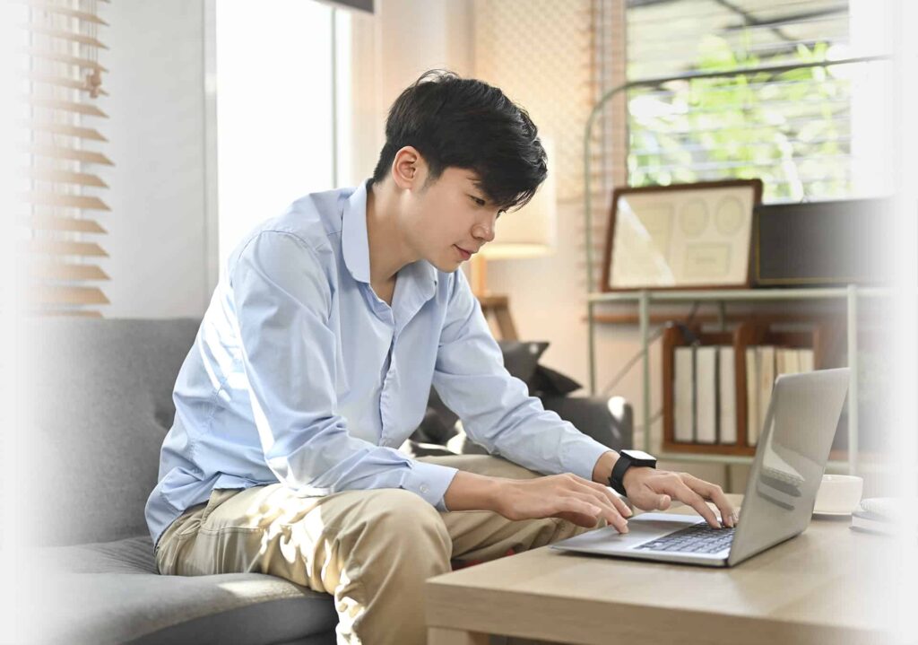 A person wearing a blue shirt and khaki pants sits on a gray sofa, typing on a laptop placed on a coffee table in a well-lit room.