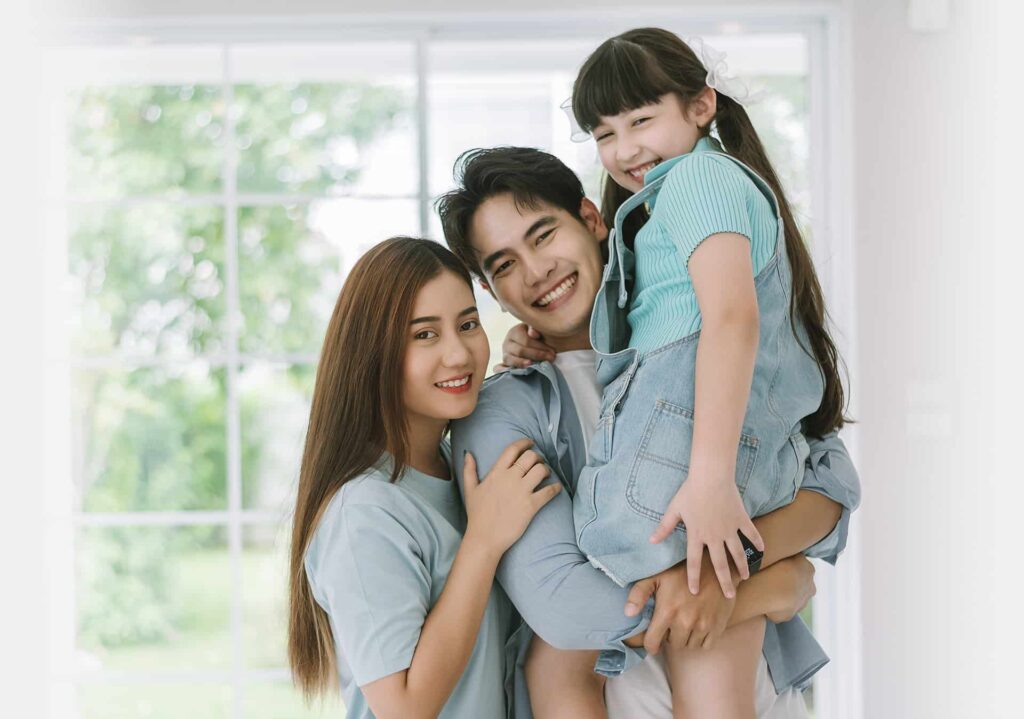 A smiling family of three poses indoors. The man, looking like a dedicated corporate executor, holds a young girl while the woman stands next to them. They are in front of a large window with a blurred green outdoor scene.