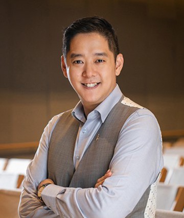 A man in a light gray shirt and vest stands with his arms crossed, smiling at the camera in an indoor setting. He appears confident, much like a seasoned professional ready to offer estate planning courses in Singapore.