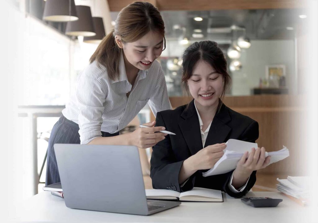 Two women at a desk in a modern office space. One is standing and pointing at documents in the other's hand. A laptop is open on the desk beside them.
