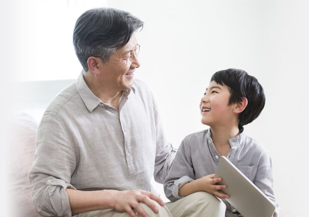 An elderly man and a young boy with a tablet sit together, smiling at each other against a bright background, illustrating the importance of ensuring proper guidance for the future through measures like Lasting Power of Attorney.