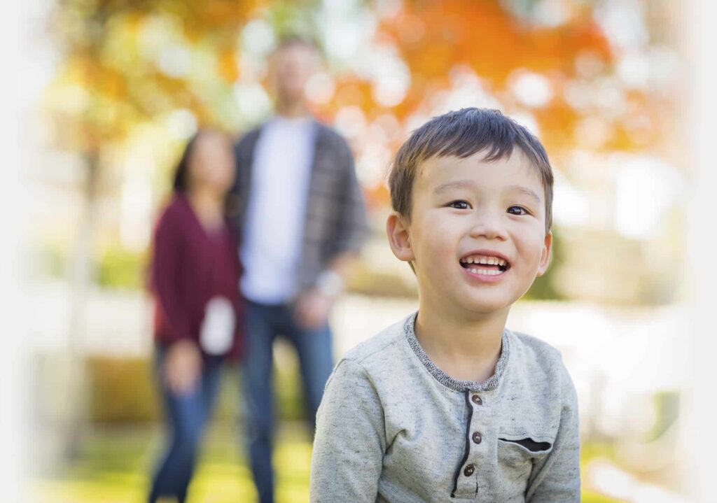 Smiling child in the foreground with two adults on standby in the blurred background on a sunny day with autumn foliage.