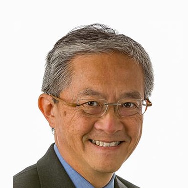 A man with short gray hair, glasses, and a suit smiles at the camera against a plain white background.