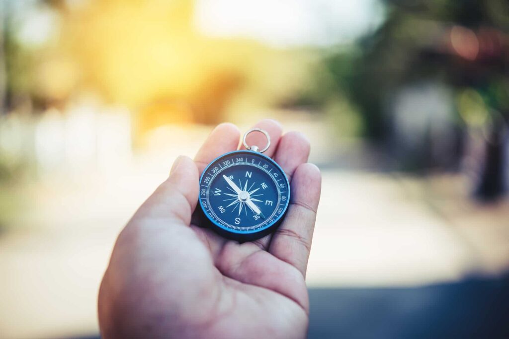 A person holds a black compass in their hand, with a blurry outdoor background visible, ready to navigate with standby trust in their direction.