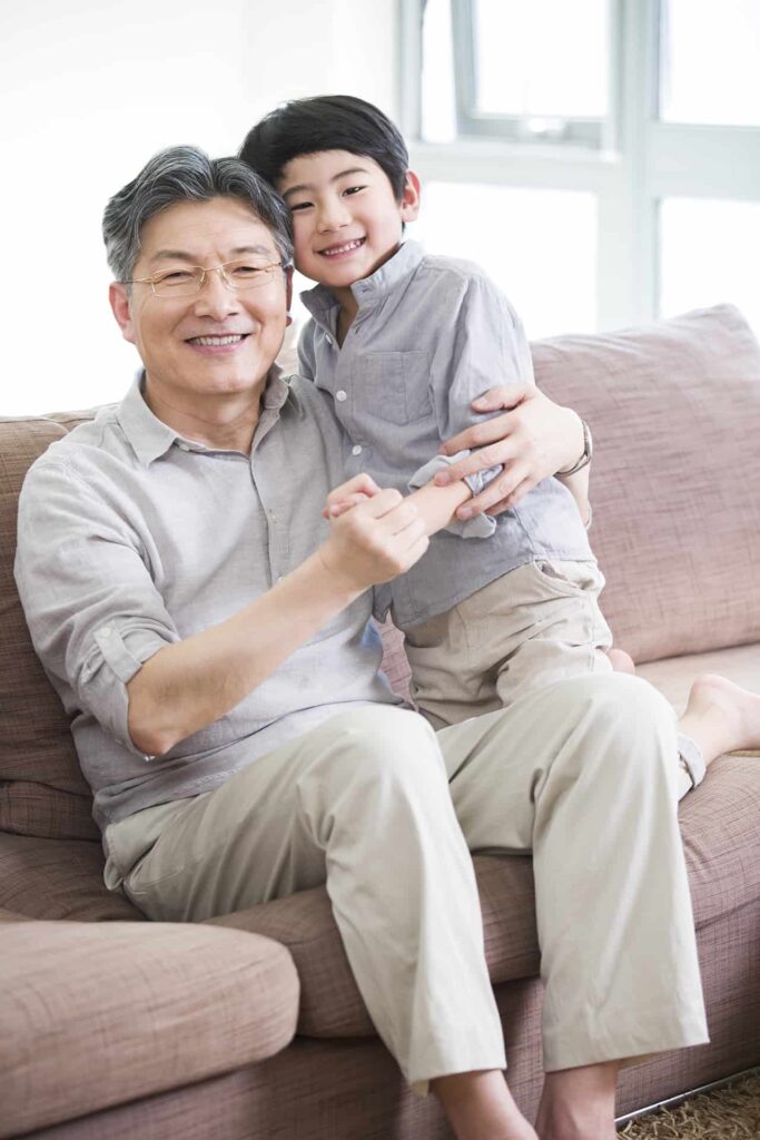 An older adult and a child, both wearing light-colored clothes, sit on a couch and smile at the camera. The older adult, who looks like they could be a corporate executor in their off-time, has glasses while the child is hugging them.