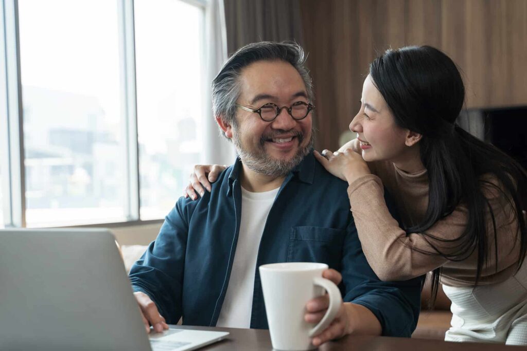 A man with glasses and a beard sits at a table with a laptop and holds a coffee mug. A woman stands beside him, smiling and resting her hands on his shoulders. They are in a bright, well-lit room.