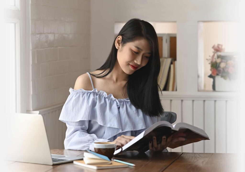 A woman with long black hair reads a book at a wooden table. A laptop, notebook, pen, and cup of coffee sit on the table beside her. She seems lost in the pages, perhaps even considering her next book purchase.