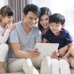 A family of four sits closely on a couch, smiling and looking at a tablet device together, perhaps exploring comprehensive will writing options to ensure their legacy protection.