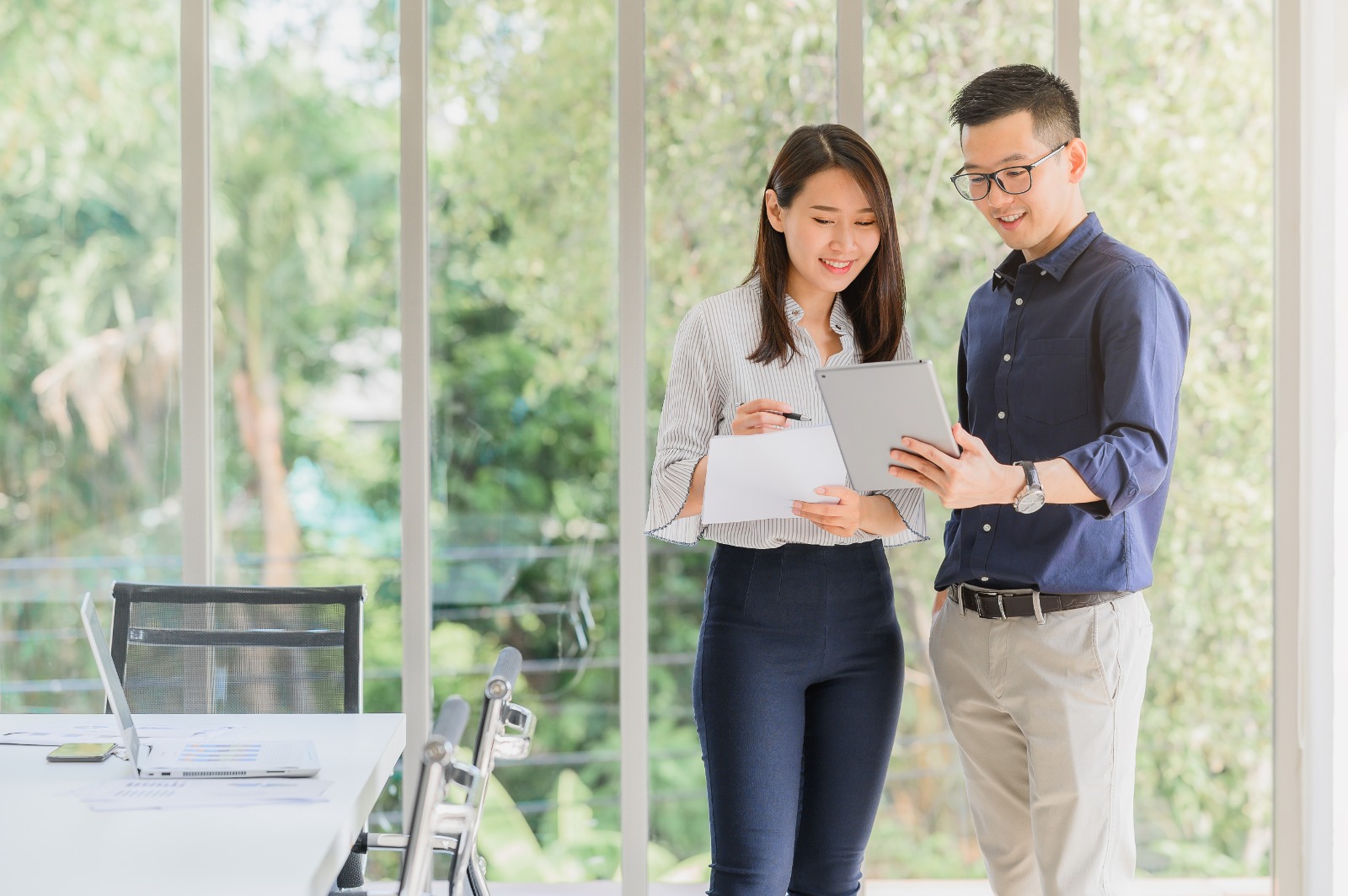 Two people in a brightly lit office space stand next to a conference table, looking at a tablet and holding documents. Their focused conversation highlights the importance of choosing the right trust company for their business in Singapore.