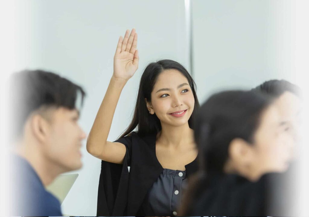 A person with long hair raises their hand in a classroom setting, smiling and looking to the side. Other students are blurred in the foreground, perhaps focused on an upcoming ESP event.