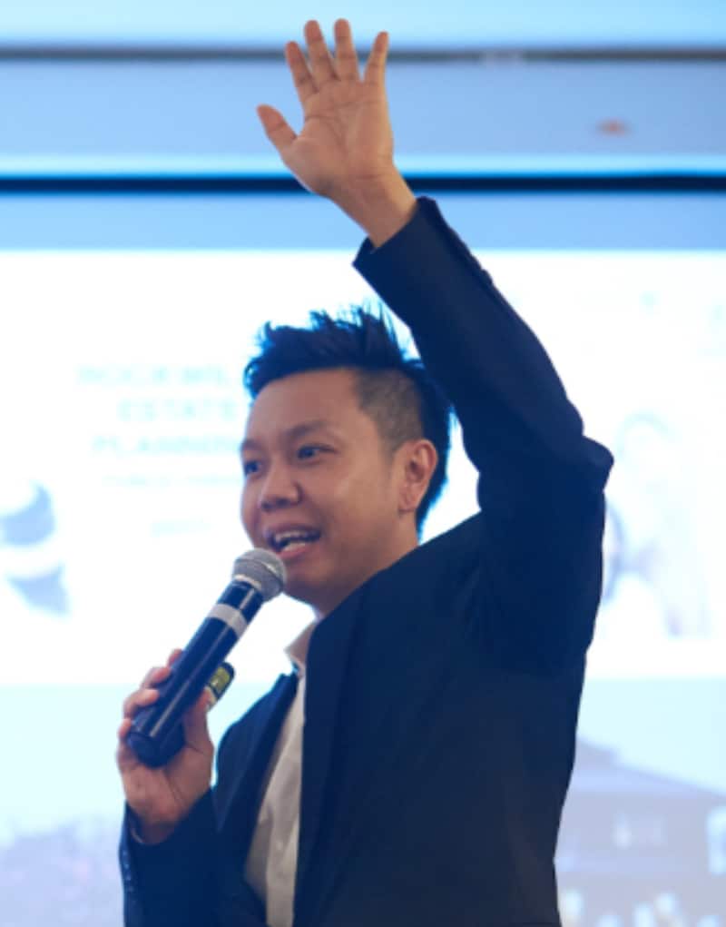 A person in a suit, possibly Eugene Soo, holds a microphone and raises one hand, appearing to speak at an Estate & Succession event. A blurred presentation slide is visible in the background.