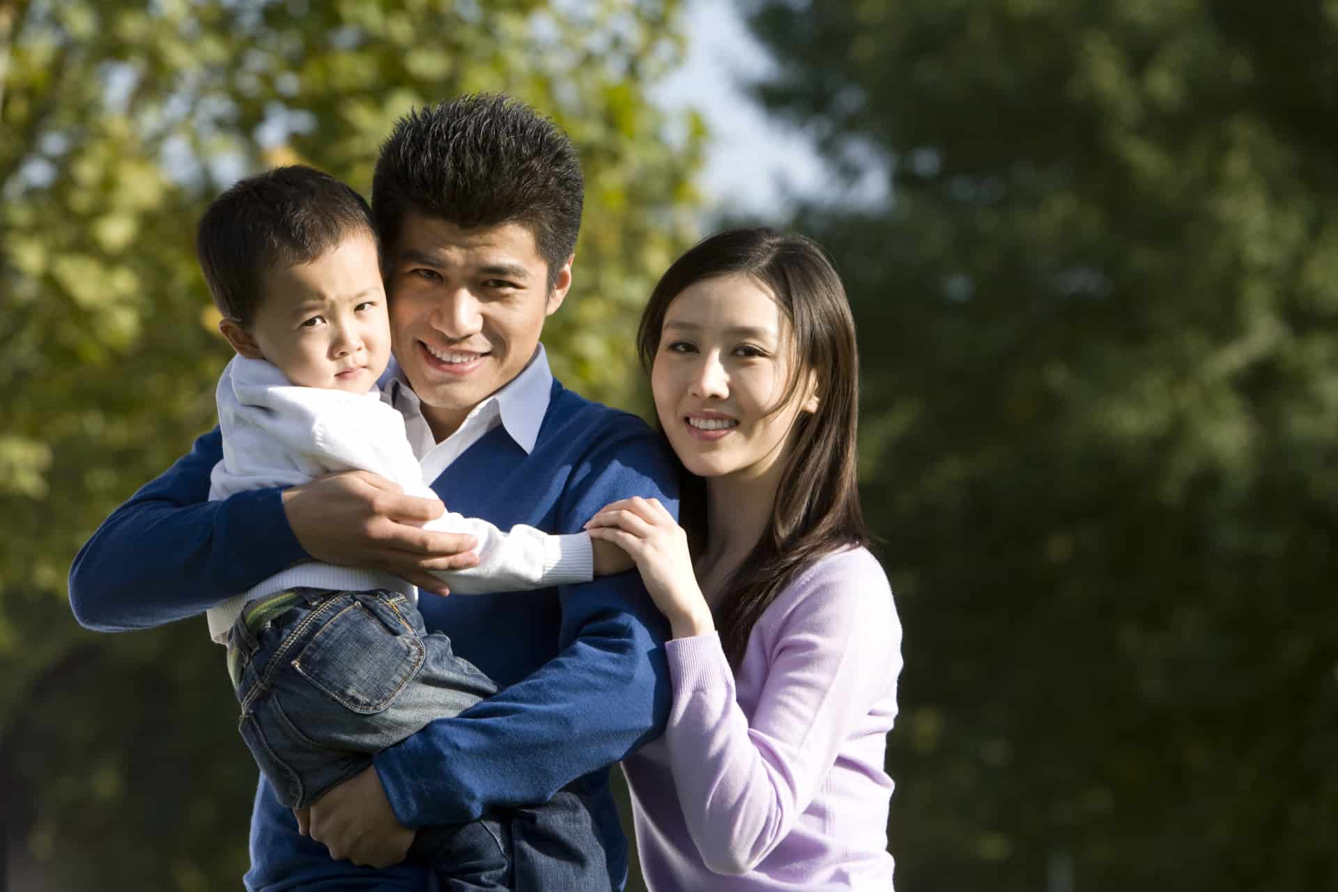 A man and woman stand outdoors, smiling at the camera, with Eugene Soo holding a young child in his arms. Trees are visible in the background.