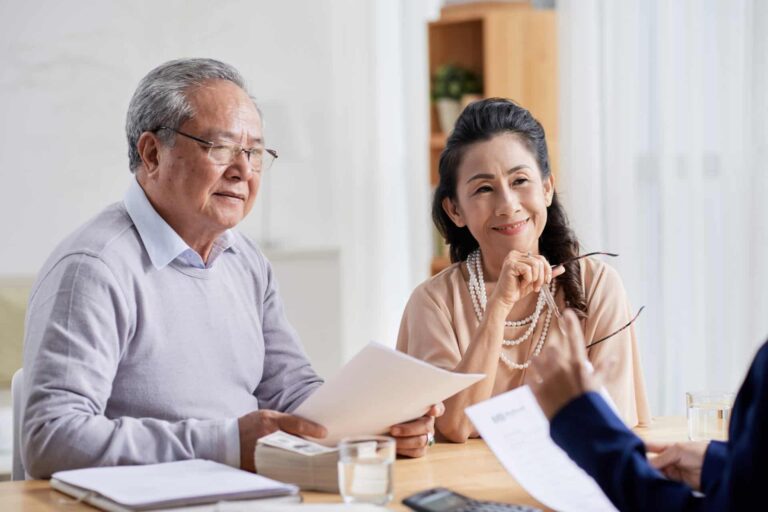 An elderly couple sits at a table reviewing documents with a person across from them. The man holds papers related to having a will, and the woman smiles while holding her glasses.